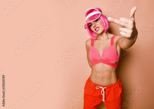 Young athletic woman with pink hair in bright sportswear standing and showing finger over yellow wall background