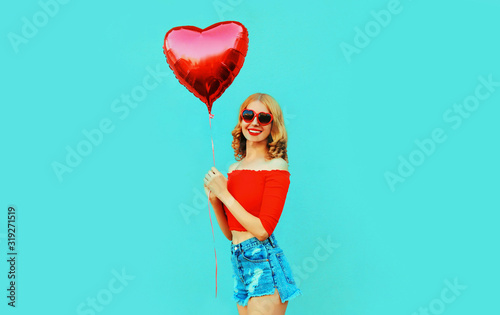 Portrait cute happy smiling woman in shorts holding red heart shaped air balloon on colorful blue background