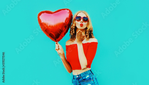 beautiful young woman sending sweet air kiss with red heart shaped balloon on colorful blue background