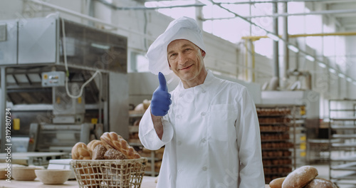In front of the camera portrait of a good looking man commercial face baker in a stylish uniform smiling large while standing beside of a working table in a bakery kitchen