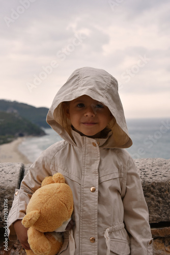 Little girl with teddy bear is wearing a raincoat jacket with hood against a stormy sky. Childhood concepts. Environmental concepts. Climate concepts. photo