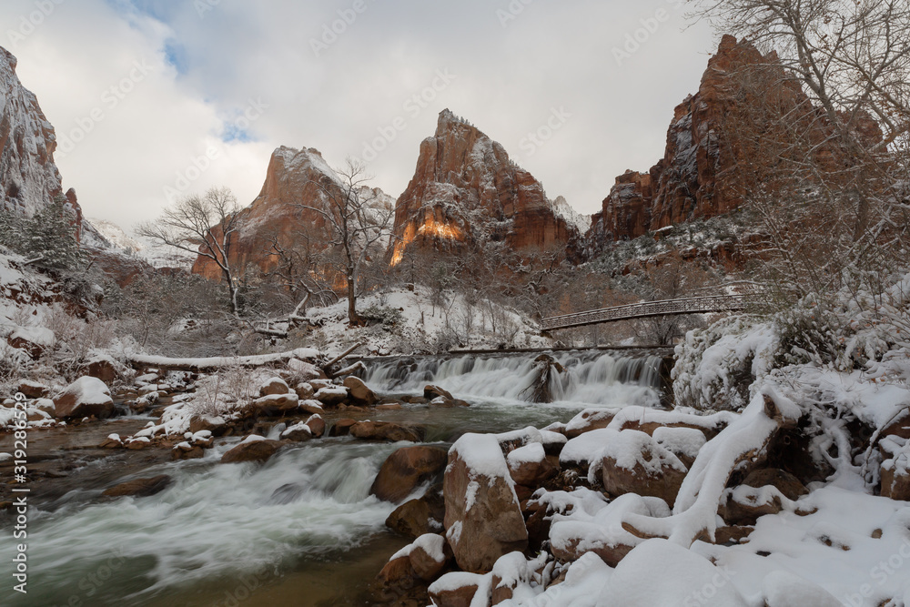 Obraz premium Snow covers the landscape in the Court of the Patriarchs in Zion National Park Utah while a small beam of sunlight breaks through the clouds to light a bit of the distant mountain.