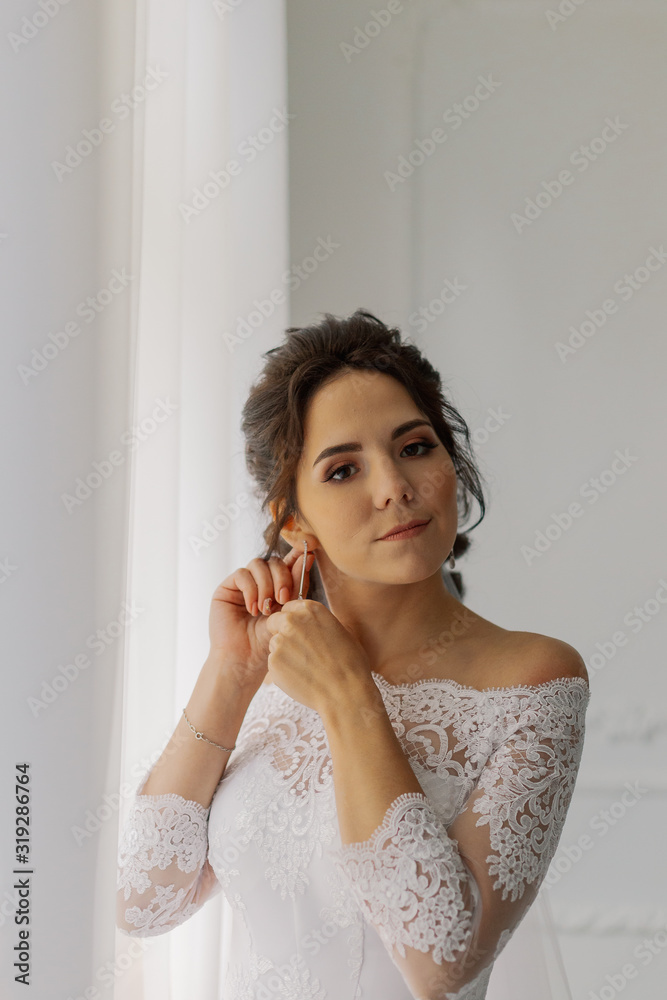 Beautiful bride puts on earrings by the window. Happy wedding day.
