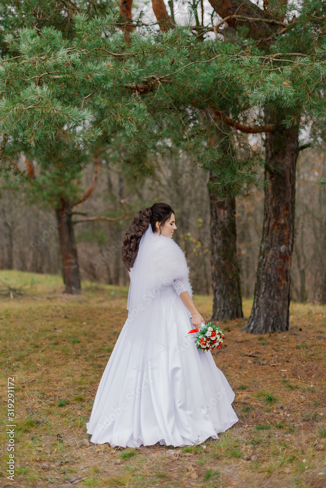 Happy bride in a beautiful wedding dress stands by the pine tree.