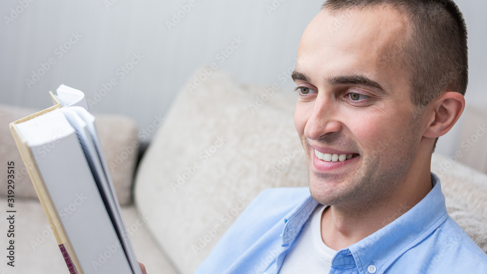 Fototapeta premium Guy reading a book sitting on sofa at home, close up, toned, 16:9