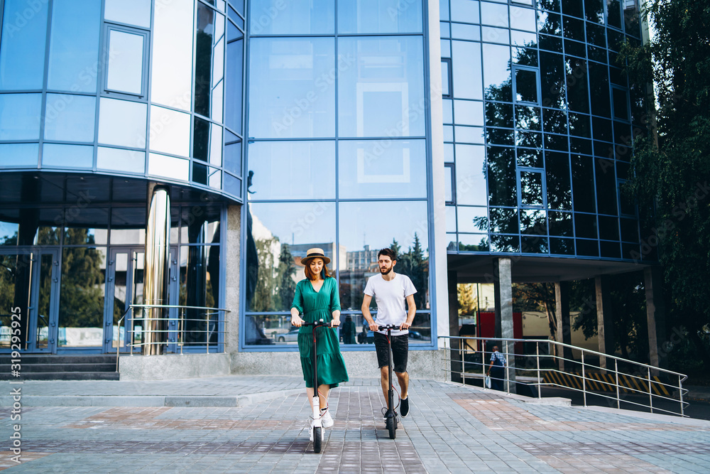Obraz premium Full length portrait of a young romantic couple with electric scooters, walking in the city. Young woman in hat and man enjoy a walk