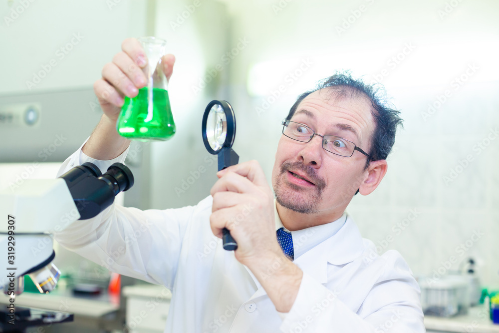 Chemist crazy. A mad scientist conducts experiments in a scientific laboratory. With surprised eyes, he looks through a magnifying glass at the green liquid in the flask.