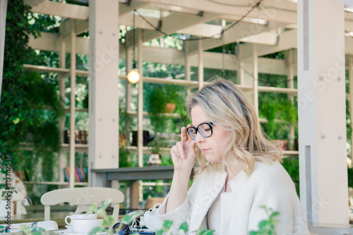 A pretty blonde in glasses and a white jacket is sitting in a summer cafe and thinking about something