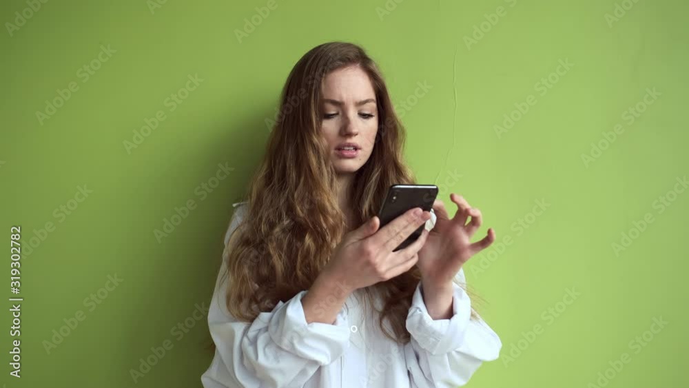Girl With Long Hair Stands Against Light Green Wall With Mobile Phone In Her Hands. Girl With Long Hair Stands Against Light Green Wall With Mobile Phone In Her Hands.