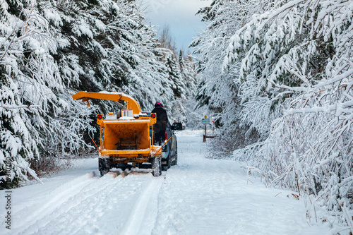 A wide angle view of a single work vehicle towing heavy machinery after a snowstorm in Quebec. Tracks are seen through thick snow with white trees and copy space