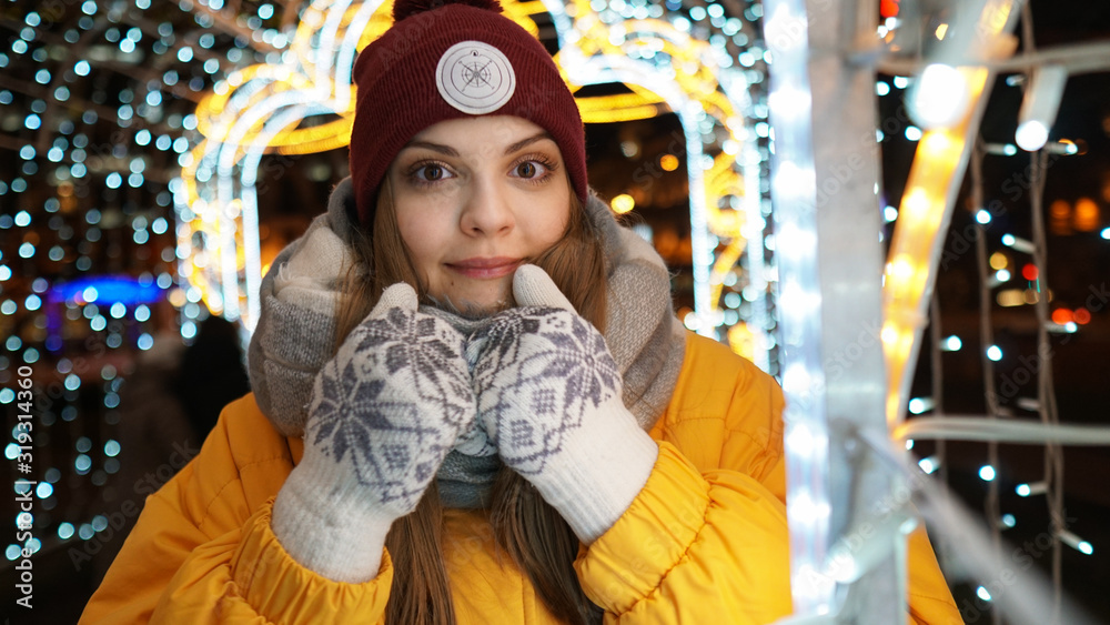 Winter portrait of young beautiful woman covering face with woolen scarf. Young woman holding scarf and looking at camera. Festive garland lights. Holiday fair. Christmas background	