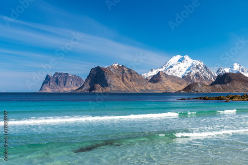 Beautiful beach during springtime on a sunny day with blue sky and silent ocean with small waves. Mountain chain with snow in the background. From Myrland beach in Lofoten - Norway. Blue clear water.