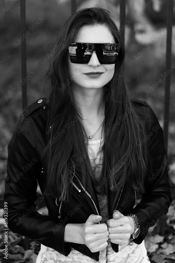 Outdoor fashion portrait of stylish young woman having fun, emotional face , laughing, Urban city street style.Girl wearing white pants,t-shirt, black leather jacket , sunglasses and dark hat Fashion.