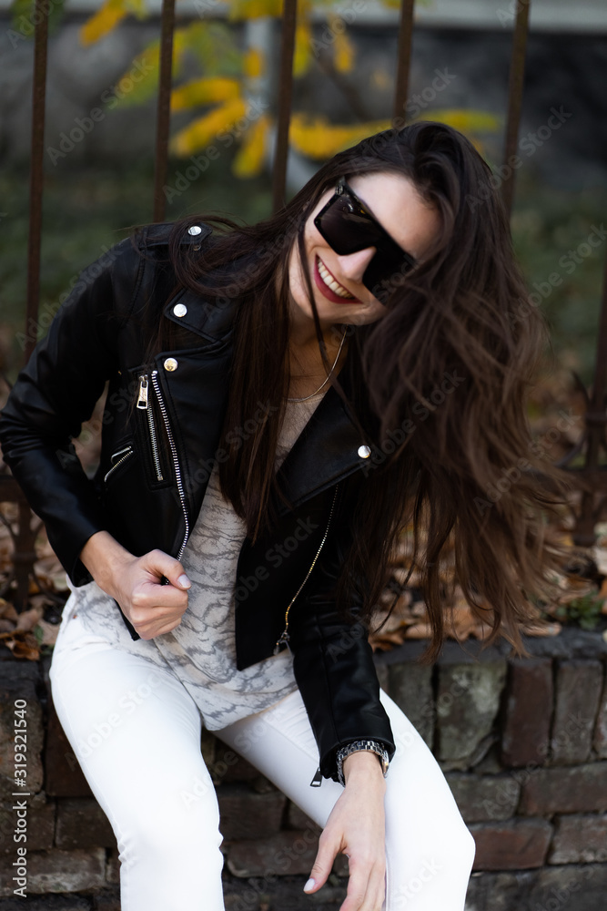 Outdoor fashion portrait of stylish young woman having fun, emotional face , laughing, Urban city street style.Girl wearing white pants,t-shirt, black leather jacket , sunglasses and dark hat Fashion.