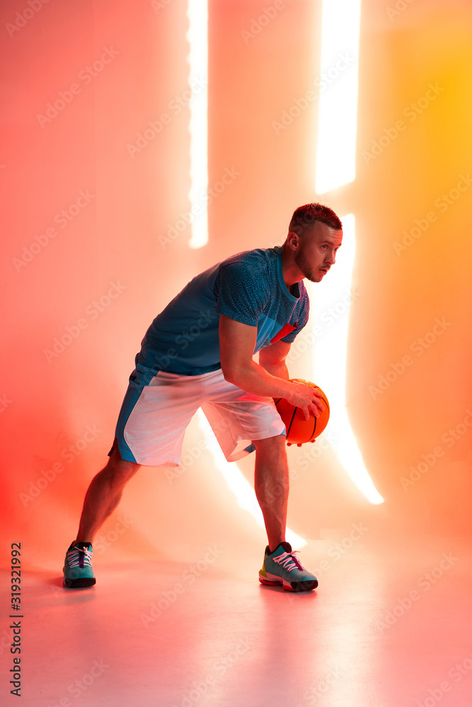 Young athletic man, basketball player dribbling with ball on red and orange background