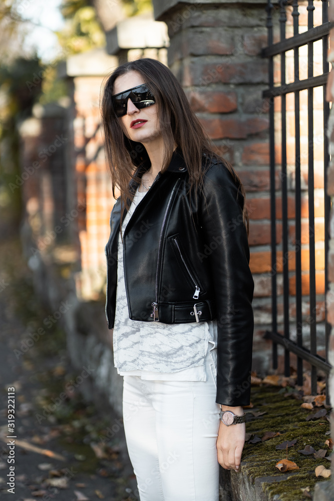 Outdoor fashion portrait of stylish young woman having fun, emotional face , laughing, Urban city street style.Girl wearing white pants,t-shirt, black leather jacket , sunglasses and dark hat Fashion.