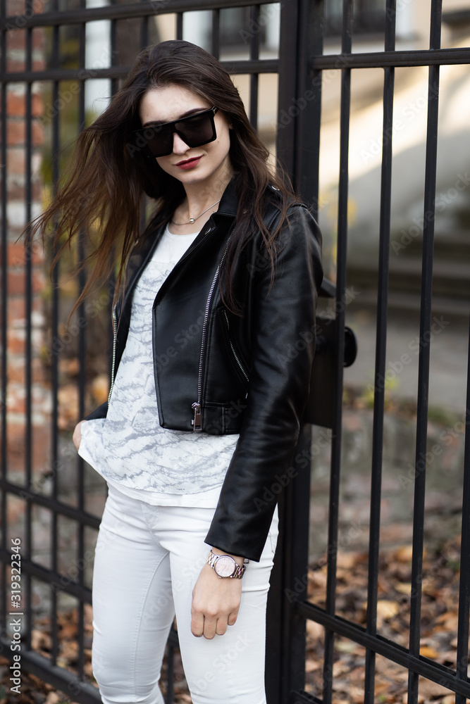 Outdoor fashion portrait of stylish young woman having fun, emotional face , laughing, Urban city street style.Girl wearing white pants,t-shirt, black leather jacket , sunglasses and dark hat Fashion.