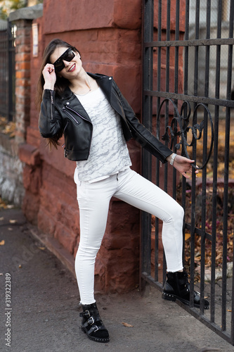 Obraz na płótnie Outdoor fashion portrait of stylish young woman having fun, emotional face , laughing, Urban city street style.Girl wearing white pants,t-shirt, black leather jacket , sunglasses and dark hat Fashion. w salonie
