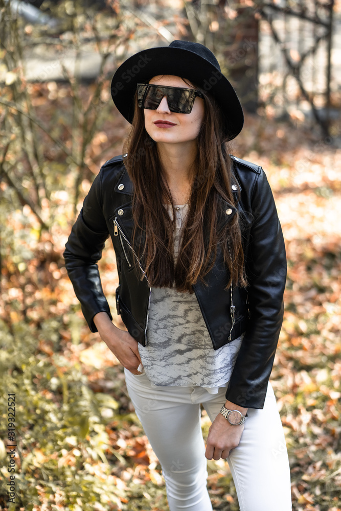 Outdoor photo of brunette lady posing on tree background in autumn day.Fashion street style portrait. Girl wearing white pants,t-shirt, black leather jacket , sunglasses and dark hat .Fashion concept.