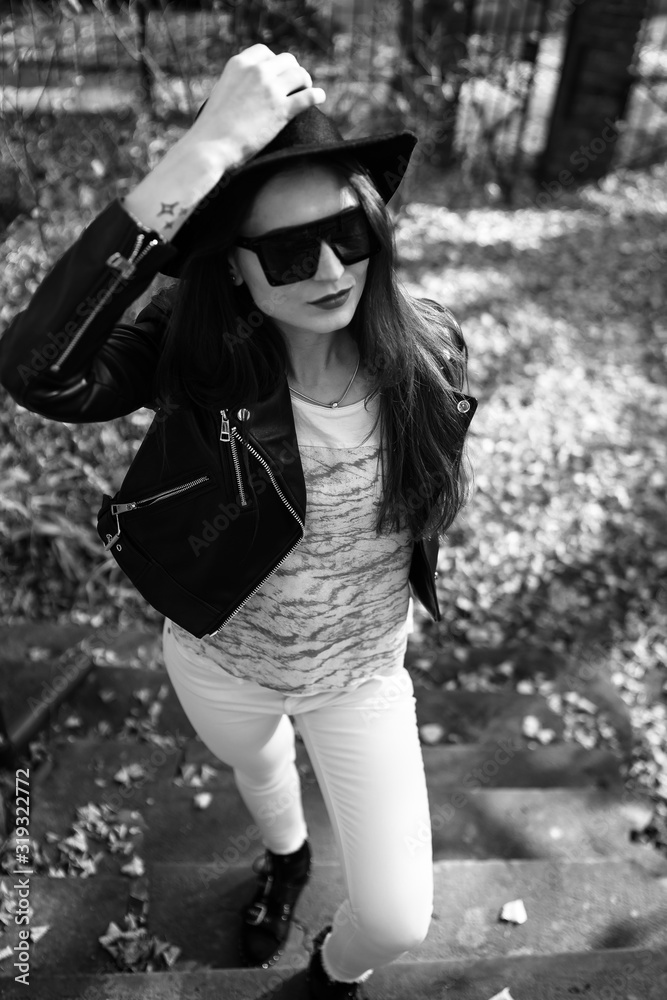 Outdoor photo of brunette lady posing on tree background in autumn day.Fashion street style portrait. Girl wearing white pants,t-shirt, black leather jacket , sunglasses and dark hat .Fashion concept.