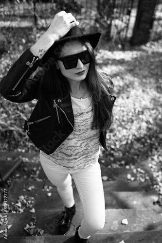 Obraz na płótnie Outdoor photo of brunette lady posing on tree background in autumn day.Fashion street style portrait. Girl wearing white pants,t-shirt, black leather jacket , sunglasses and dark hat .Fashion concept. w salonie