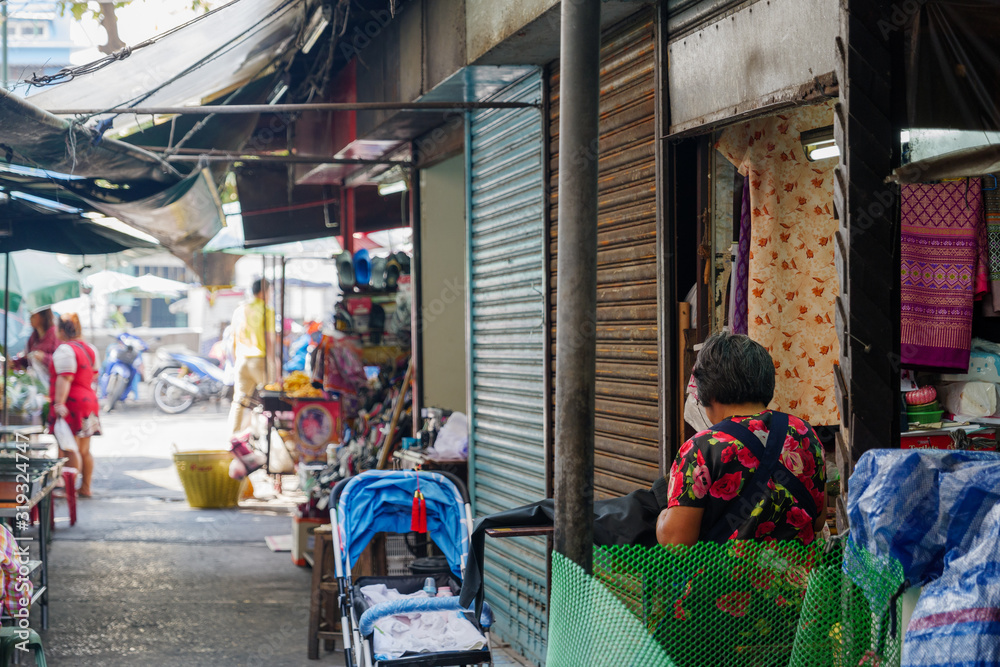 Woman sit in front of tailor shop inside old mess stall with some closing shutter doors in small narrow alley at open air market in Bangkok, Thailand.