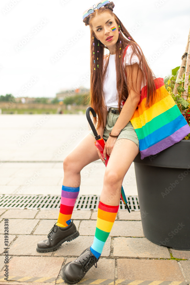 Stylish bisexual girl with lgbt rainbow on her face posing at city square with colorful umbrella and bag.