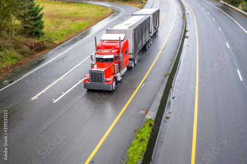 Red classic big rig semi truck with spoiler running on the turning wet road transporting two semi trailers with commercial cargo