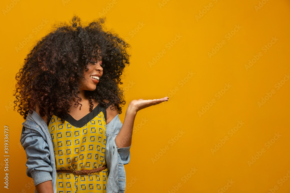 Cheerful Afro woman points away on copy space, discusses amazing promo, gives way or direction, wears yellow warm sweater, has pleasant smile, feels optimistic, isolated over yellow background. Cheerful Afro woman points away on copy space, discusses amazing promo, gives way or direction, wears yellow warm sweater, has pleasant smile, feels optimistic, isolated over yellow background.