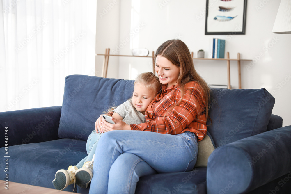 Young mother and daughter with mobile phone sitting on sofa at home