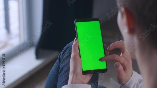 Girl with short hair sitting home in armchair and fingers display green screen cellphone tapping scrolling message. Woman with telephone on a winter. Chroma key mobile phone greenscreen technology.