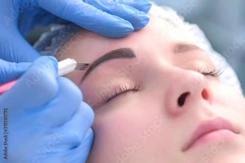 Beautician is painting shaping contour using manual tool for tattoo  woman s face closeup. Cosmetologist making eyebrows microblading procedure in beauty salon for girl. Beauty industry concept.