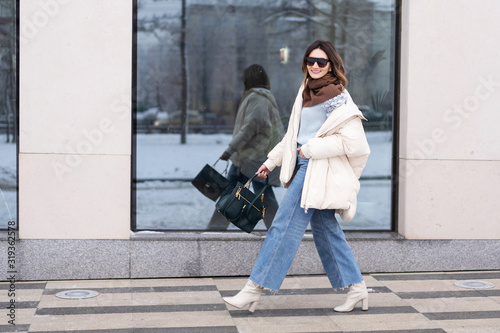 European model girl in a beige oversized down jacket, knitted sweater, jeans flared with a handbag and in glasses is walking along the street near glass windows. Life style