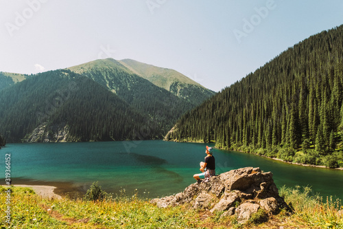 Kazakhstan, Kolsai Lake. The second Kolsai Lake in the mountains among pine forests in the sunlight. Turquoise water and yellow grass A blonde girl in shorts is standing by the lake