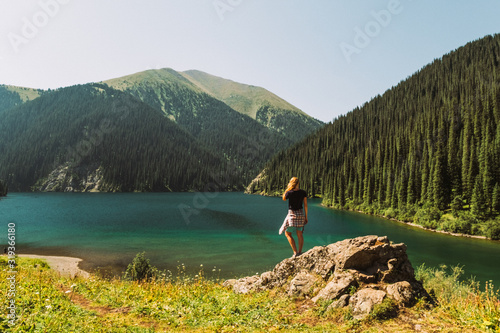 Kazakhstan, Kolsai Lake. The second Kolsai Lake in the mountains among pine forests in the sunlight. Turquoise water and yellow grass A blonde girl in shorts is standing by the lake