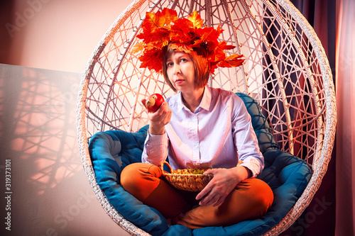 Ugly girl in a white shirt, red jeans and with a wreath of yellow autumn maple leaveson and with apple in a white wicker hanging chair photo