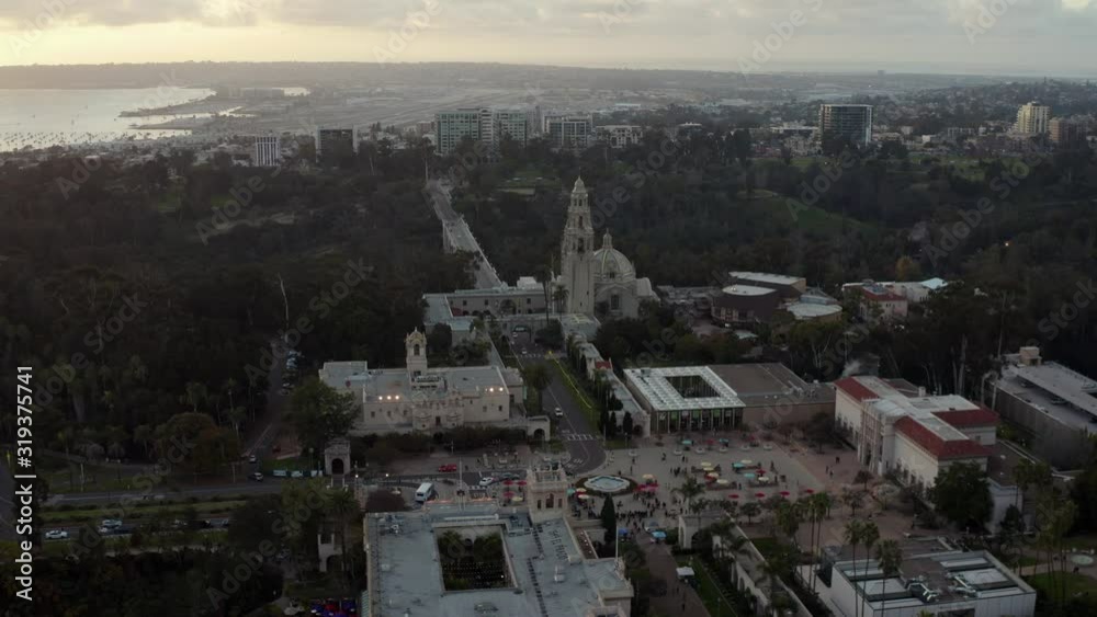 Aerial drone footage of the iconic buildings and towers of Balboa Park during a beautiful Sunset with the San Diego, California skyline is the background. 