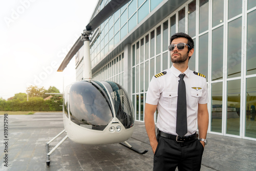 Portrait of handsome commercial pilot in captain white uniform standing and looking smart near small private helicopter on a landing point with private jet terminal at international airport. photo