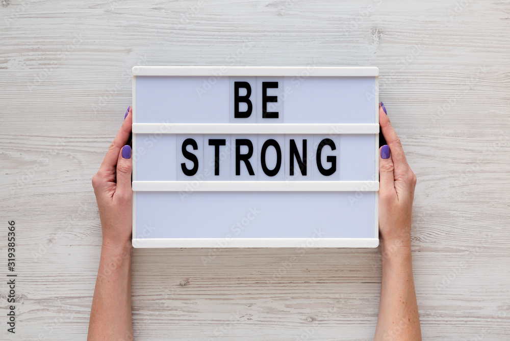 Female hands hold a lightbox with 'Be strong' words over white wooden surface, top view. Overhead, from above, flat lay. Female hands hold a lightbox with 'Be strong' words over white wooden surface, top view. Overhead, from above, flat lay.