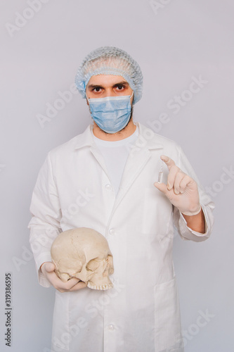 The doctor holds a skull and an ampoule with a vaccine in his hands. A young guy in a white coat, in a medical mask and gloves on a white background. Pathologist medical worker. Disease mortality conc