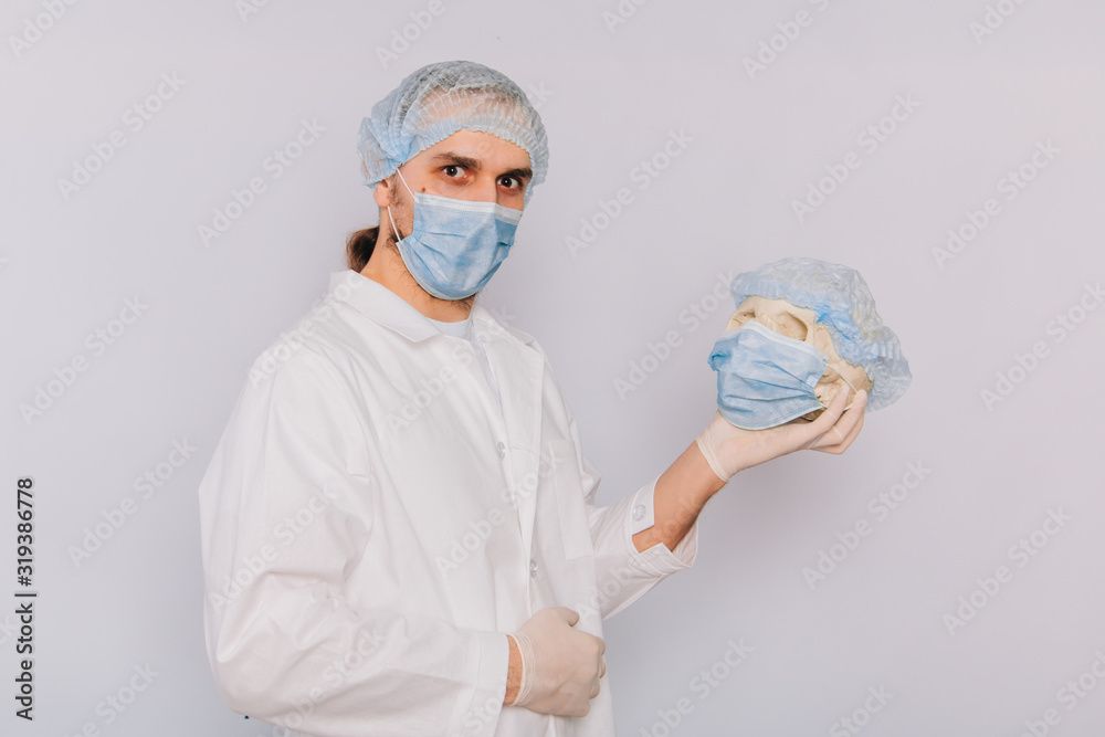The doctor holds a skull in his hands. A young guy in a white coat, in a medical mask and gloves on a white background. Skull meditsinskiy worker. Mortality at work concept