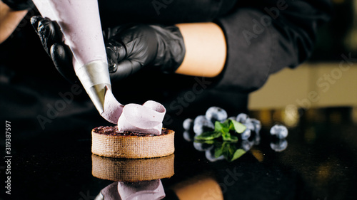 Pastry chef decorates biscuit with purple cream from pastry bag, close-up. Preparation of blueberry cake at commercial bakery with piping bag