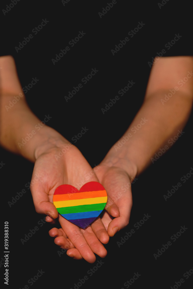 Female arms holding the heart coloured in LGBT pride colours on the dark background. Concept of the International Day Against Homophobia concept, sexual equality, feminism, social safety