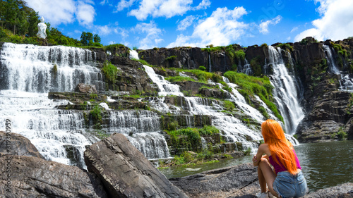 Beautiful young red haird girl sitting near amazing  waterfall in Da Lat city Vietnam.Traveler girl with long red hair looking to beautiful Pongour  waterfall. photo