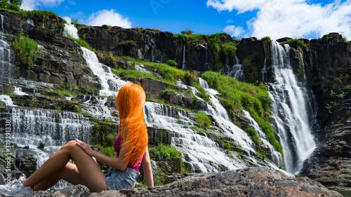 Beautiful young red haird girl sitting near amazing waterfall in Da Lat city Vietnam.Traveler girl with long red hair looking to beautiful Pongour waterfall.