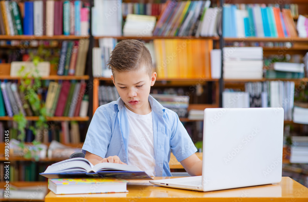 A boy with glasses in a library at a table works on a book, an open laptop is standing nearby A boy with glasses in a library at a table works on a book, an open laptop is standing nearby