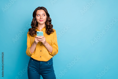 Photo of pretty wavy lady holding telephone hands thinking over creative post text idea looking side empty space wear yellow shirt trousers isolated blue color background © deagreez