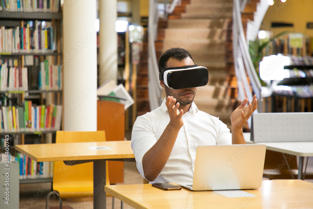Obraz premium Focused guy with VR headset sitting in library. Serious African American man experiencing new VR glasses. Technology concept