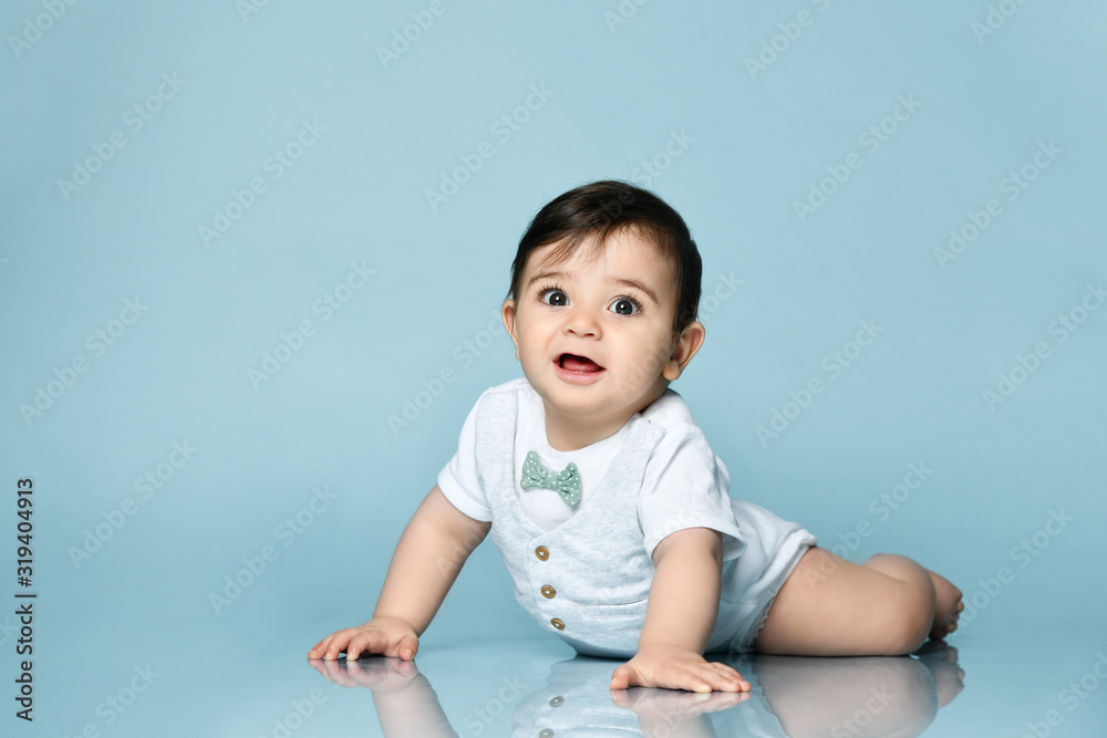 Baby boy in white bodysuit as a vest with bow-tie, barefoot. He is laying on the floor against blue background. Close up