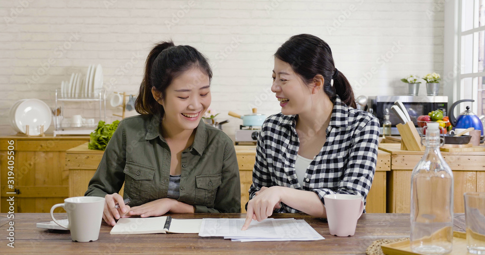 Happy young female friends calculating bills at home kitchen. ladies smiling and talking while sitting at dining table analyzing finance. asian chinese women roommates discussing about spend money. Happy young female friends calculating bills at home kitchen. ladies smiling and talking while sitting at dining table analyzing finance. asian chinese women roommates discussing about spend money.